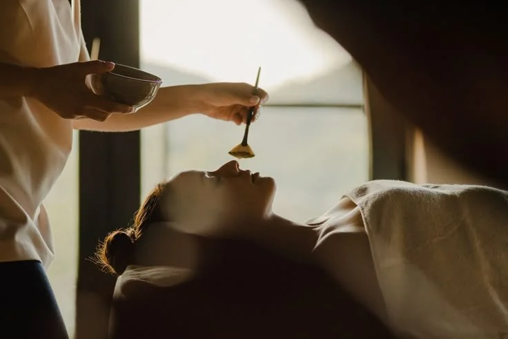 A woman receiving a facial treatment, lying on a bed with her head supported, while an esthetician applies a mask or cream to her face with a brush in a softly lit room.