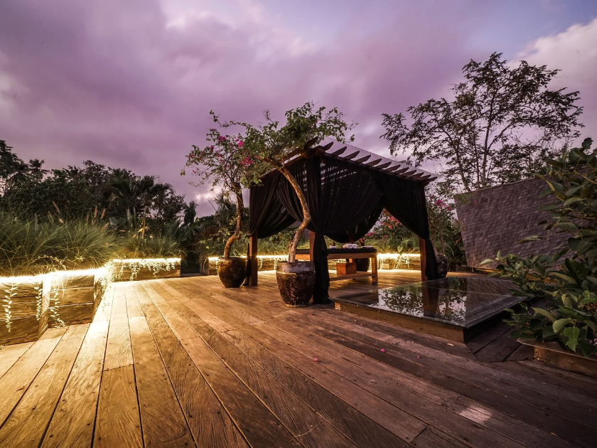 A wooden deck with a seating area under a canopy with black fabric curtains, surrounded by potted plants, fairy lights along the edge, and trees with a purple sky overhead at dusk.