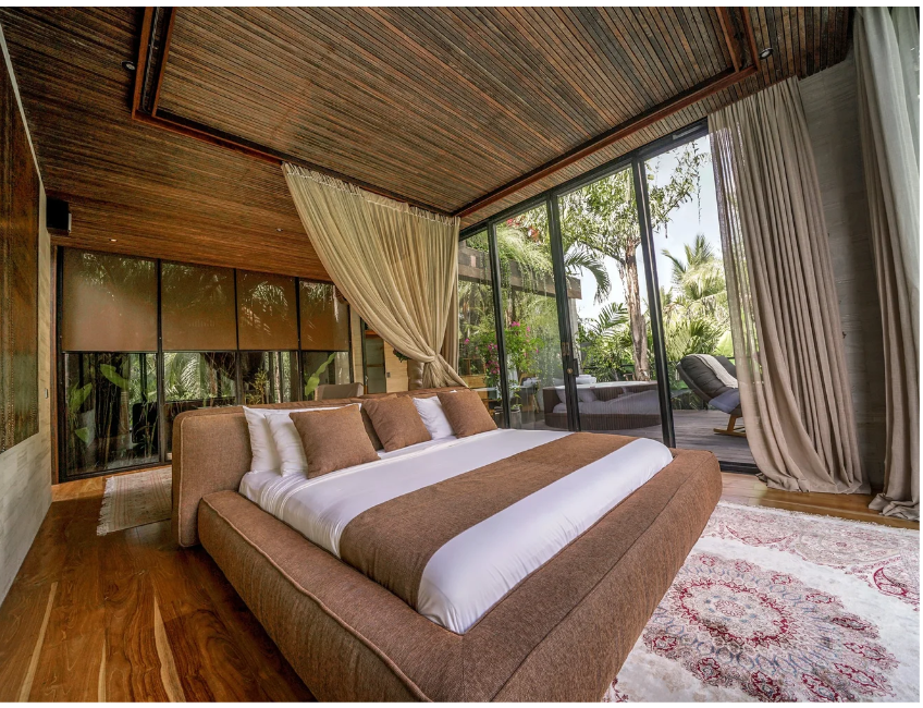 A bedroom with a brown upholstered bed, white and brown pillows, and a brown bed runner, positioned against large glass sliding doors opening to an outdoor patio with lounge chairs and lush tropical plants.