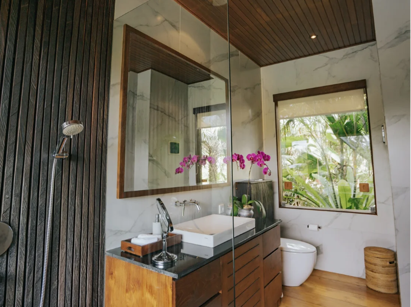 Modern bathroom with marble walls, wooden accents, and a large window overlooking greenery. Features a square sink, orchid flowers, and a wooden vanity.