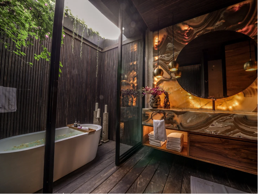 A luxurious bathroom with an outdoor bathtub on a wooden deck, separated by glass walls, with a dark wood and marble patterned interior, illuminated warm lighting, flowers, and neatly folded towels.