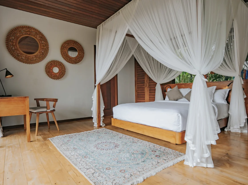 A bedroom with a four-poster bed draped with white curtains, a wooden desk and chair, decorative wall mirrors, a patterned area rug, and a wooden ceiling.