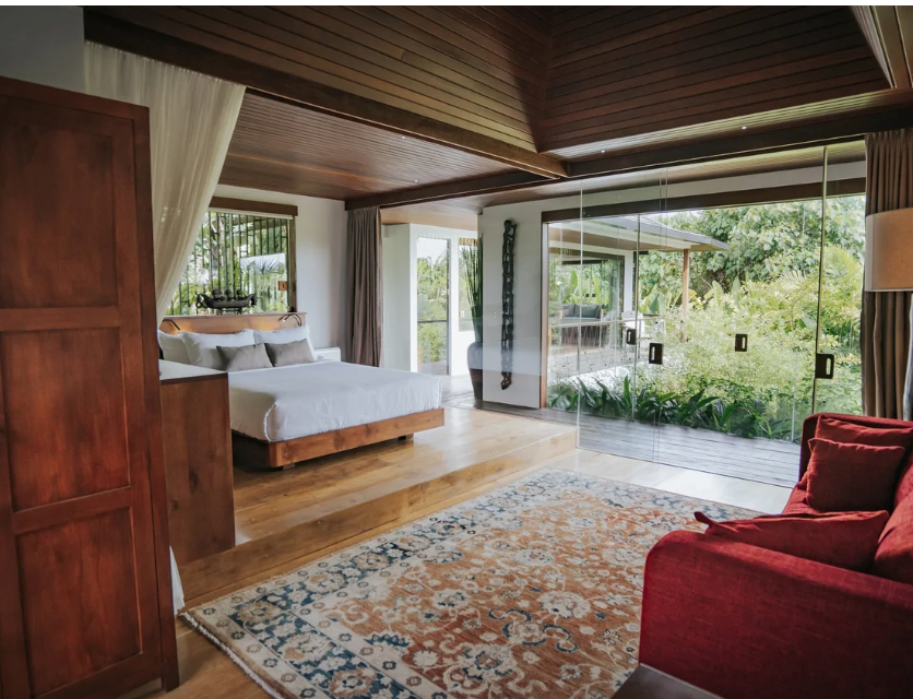 A cozy bedroom with a wooden ceiling, white walls, and wooden flooring. There is a bed with white linens and pillows, a large mirror reflecting greenery outside, and a red armchair. Sliding glass doors lead to a balcony with outdoor seating and lush 