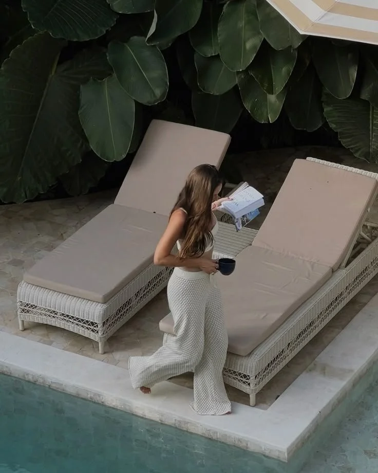 A woman with long brown hair, wearing white patterned pants and a sleeveless top, standing by a poolside on a beige lounge chair, holding a black mug, reading a book, with large green tropical leaves overhead.