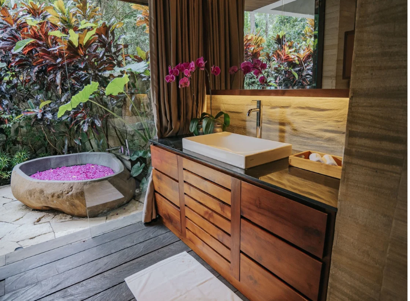 Indoor bathroom with wooden cabinetry, a white rectangular sink, a pink orchid plant, a wooden tray with toiletries, and an outdoor garden with a stone bathtub filled with pink flower petals.