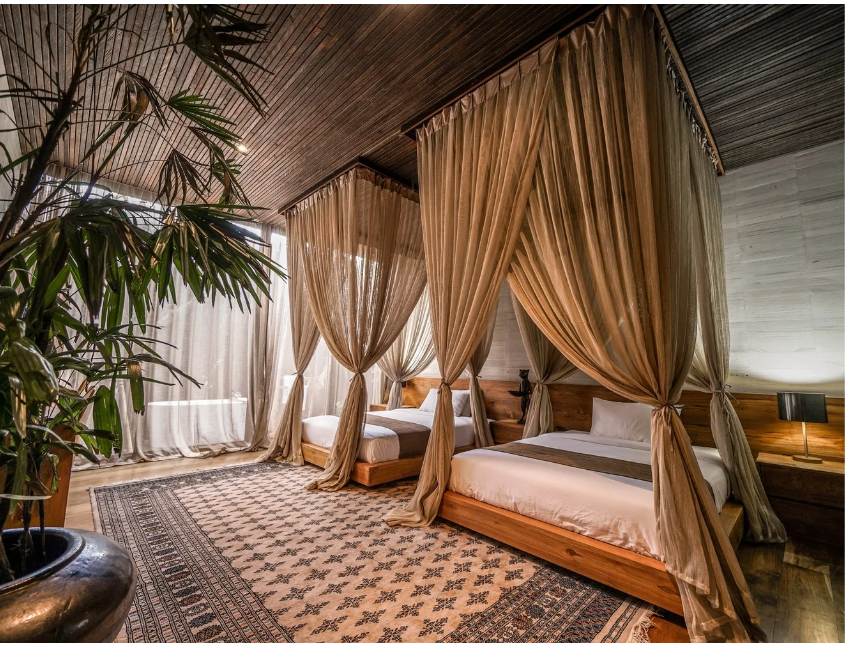 A luxurious bedroom with two canopy beds draped in beige curtains, a patterned area rug, large windows with sheer curtains, and a potted plant in the corner.
