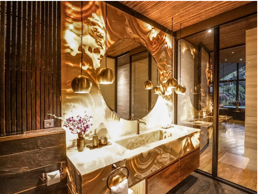 Luxury bathroom with backlit onyx countertop, round mirror, pendant lights, and wooden accents.