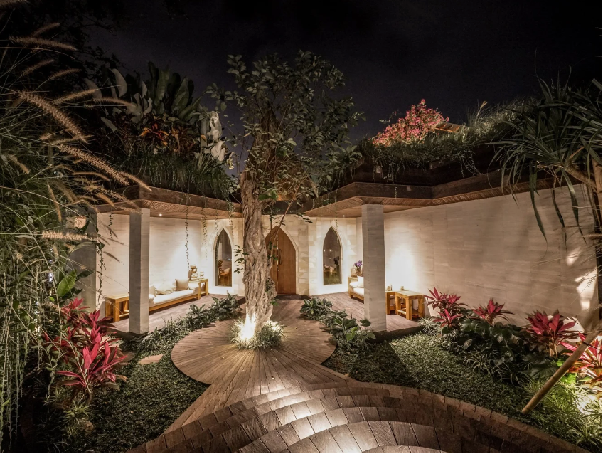 Nighttime view of a modern outdoor garden with a central tree surrounded by wooden pathways, lush plants, and seating areas with benches and pillows, illuminated by soft lighting.