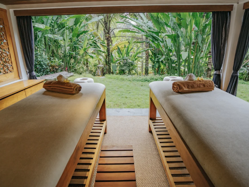 Two massage tables facing a large open window with lush green tropical plants outside, each with a rolled towel on top and a brown blanket, in a serene spa or wellness room.