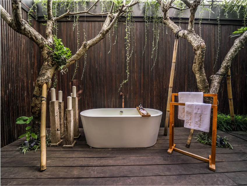 Outdoor bathtub in a natural setting with wooden fencing, trees, and plants, accompanied by a towel rack with towels and a tray with drinks.
