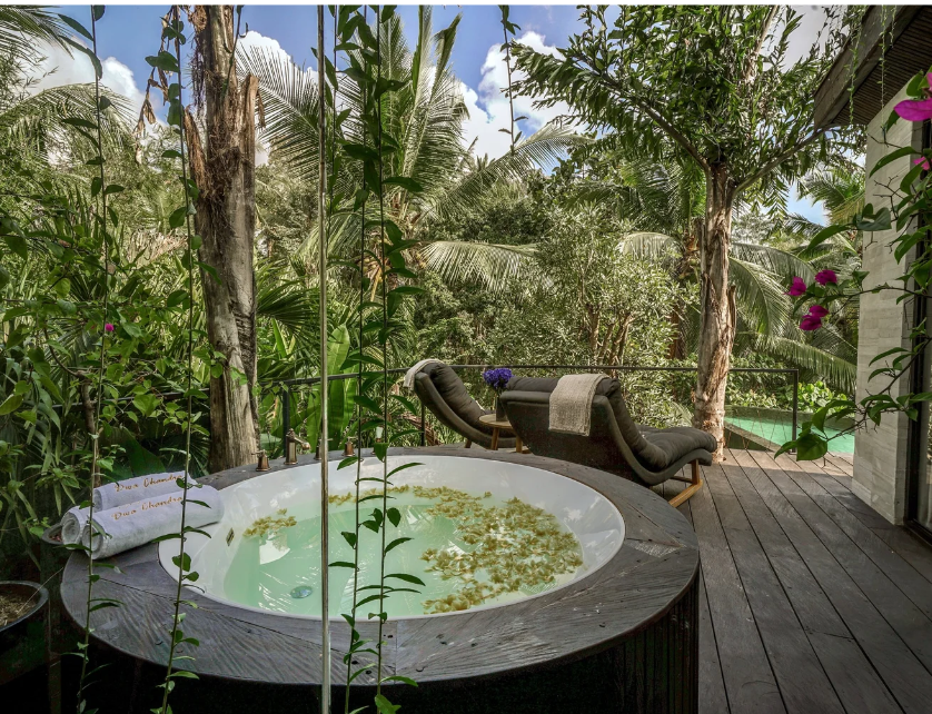 Outdoor balcony featuring a halo-shaped hot tub with floating leaves, two lounge chairs with towels and a blanket, surrounded by lush green tropical plants and trees, with a view of dense jungle foliage and a bright blue sky.