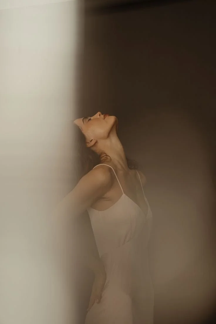 A woman in a white dress, standing with her head tilted back and eyes closed, with her back against a wall.