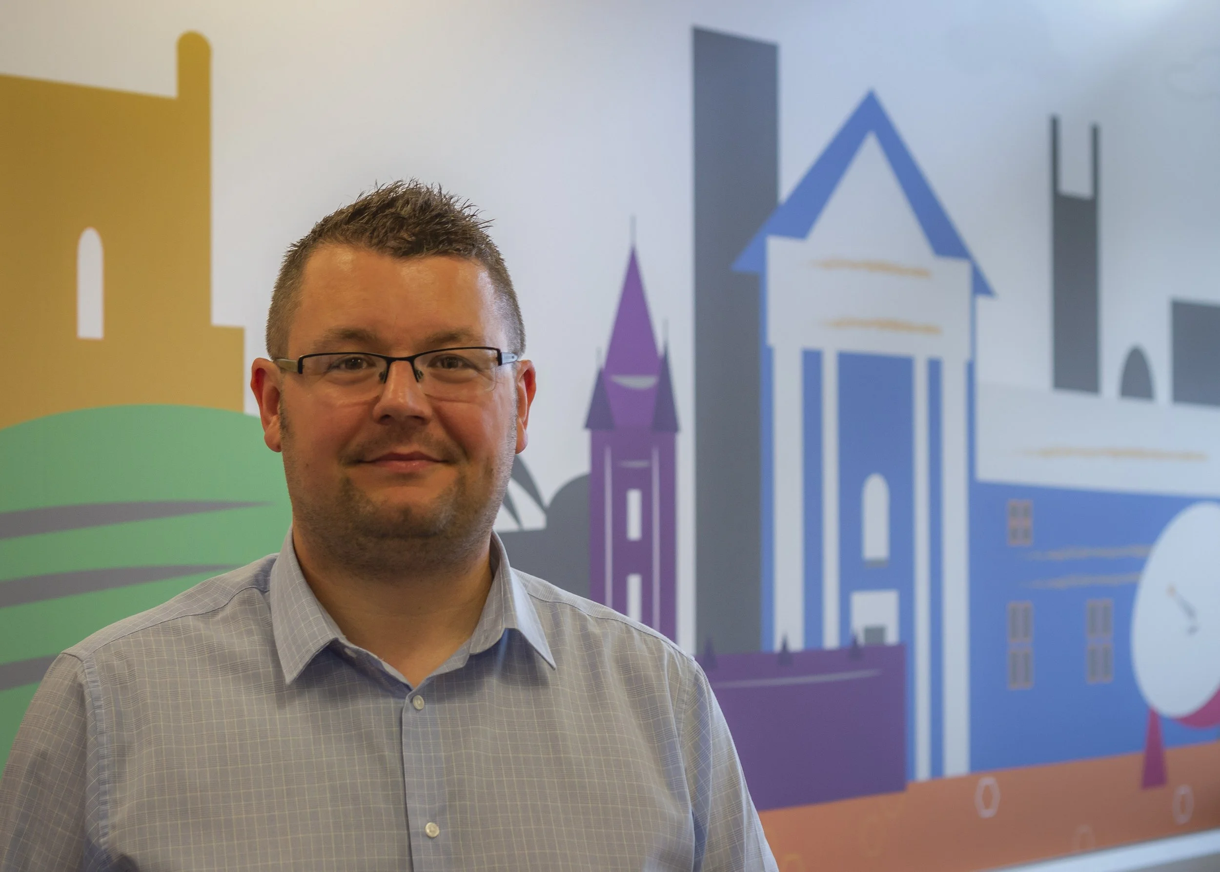 A man with glasses smiling, standing in front of a colorful mural featuring stylized illustrations of buildings and a cityscape.