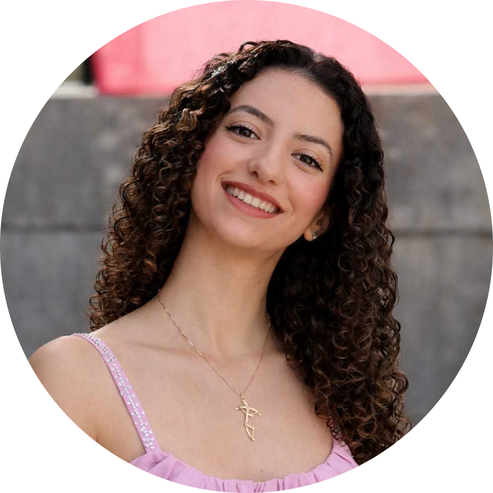 A young woman with long, curly brown hair, fair skin, and a bright smile posing outdoors in front of a concrete wall with a pink railing.
