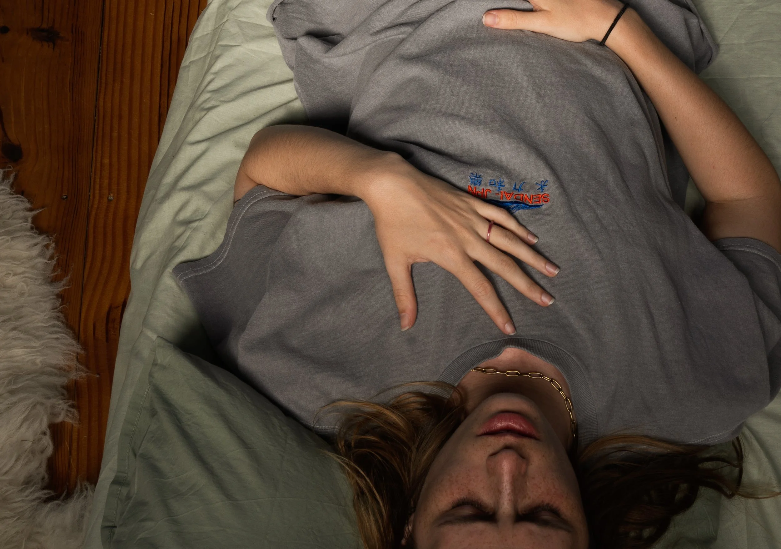 A person lying on a bed with their hand on their stomach, wearing a gray t-shirt with text and a gold chain necklace, surrounded by a wooden headboard and a furry white object near the bottom left corner.