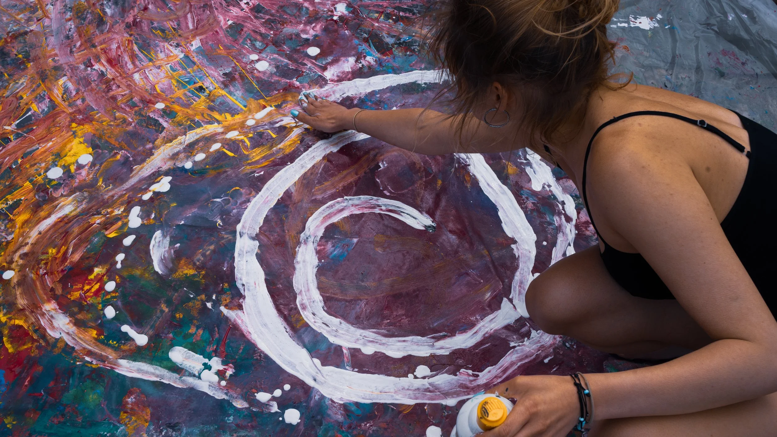 A woman with brown curly hair, earrings, and a black tank top is kneeling on the floor, working on an abstract painting with vibrant colors including purple, yellow, white, and orange, using white paint from a bottle.
