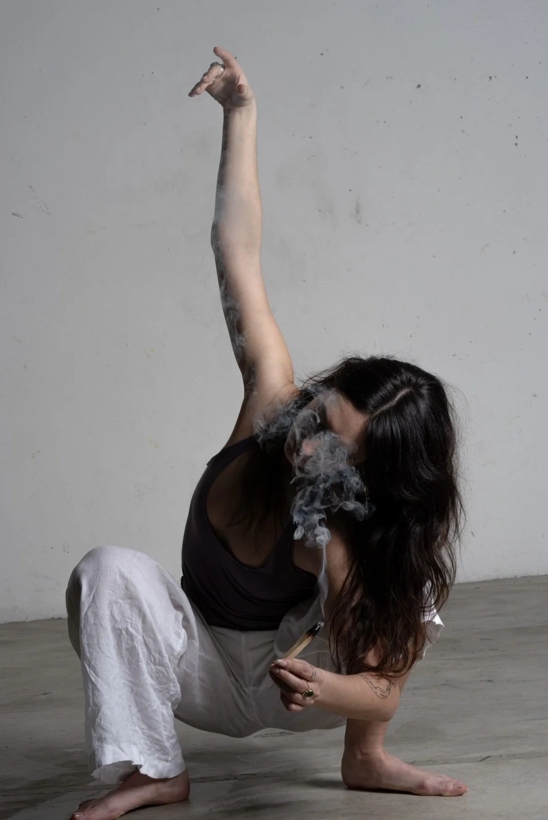 A woman with long dark hair is in a yoga pose on a wooden floor, with her left arm extended upward and smoke coming from a smoking stick in her right hand, against a plain gray wall background.