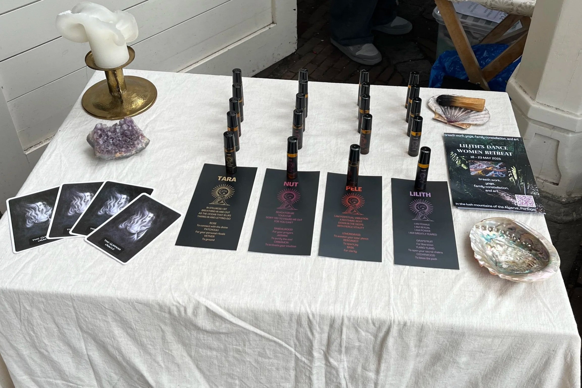 A table covered with a white cloth displays tarot cards, colored pens, a pamphlet for a women's retreat, a shell with a wooden stick, a purple mineral cluster, a candle holder with a candle, and decorative shells. In the background are partial views of people, one standing with shoes visible, and objects on the ground.