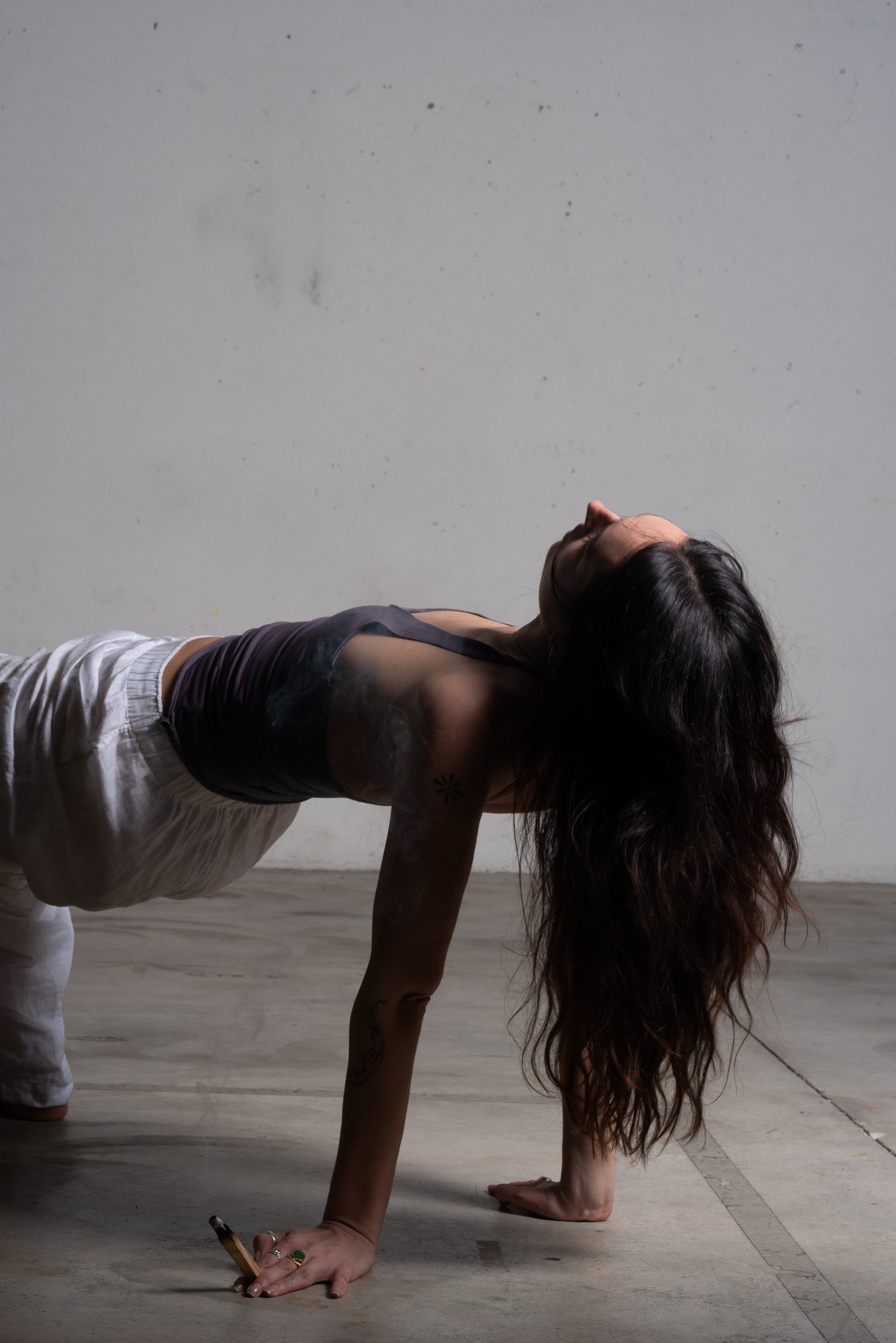 A woman with long, wavy hair doing a yoga pose on a wooden floor against a plain white wall. She is supporting herself with one hand and foot, with her other hand holding a pen, and her head tilted back.