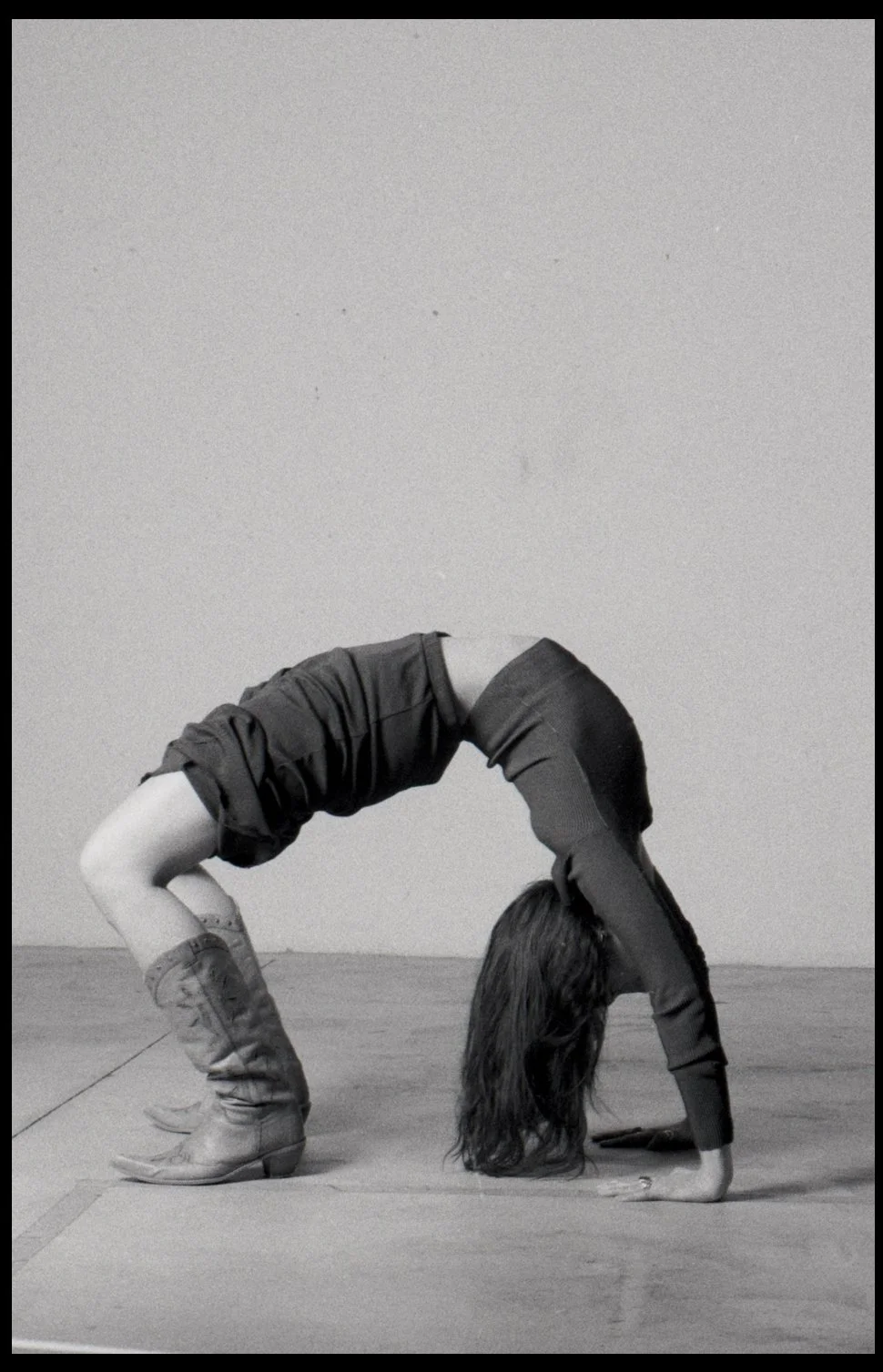 A woman in bootcut jeans and a long-sleeved shirt performing a backbend yoga pose indoors against a plain wall.