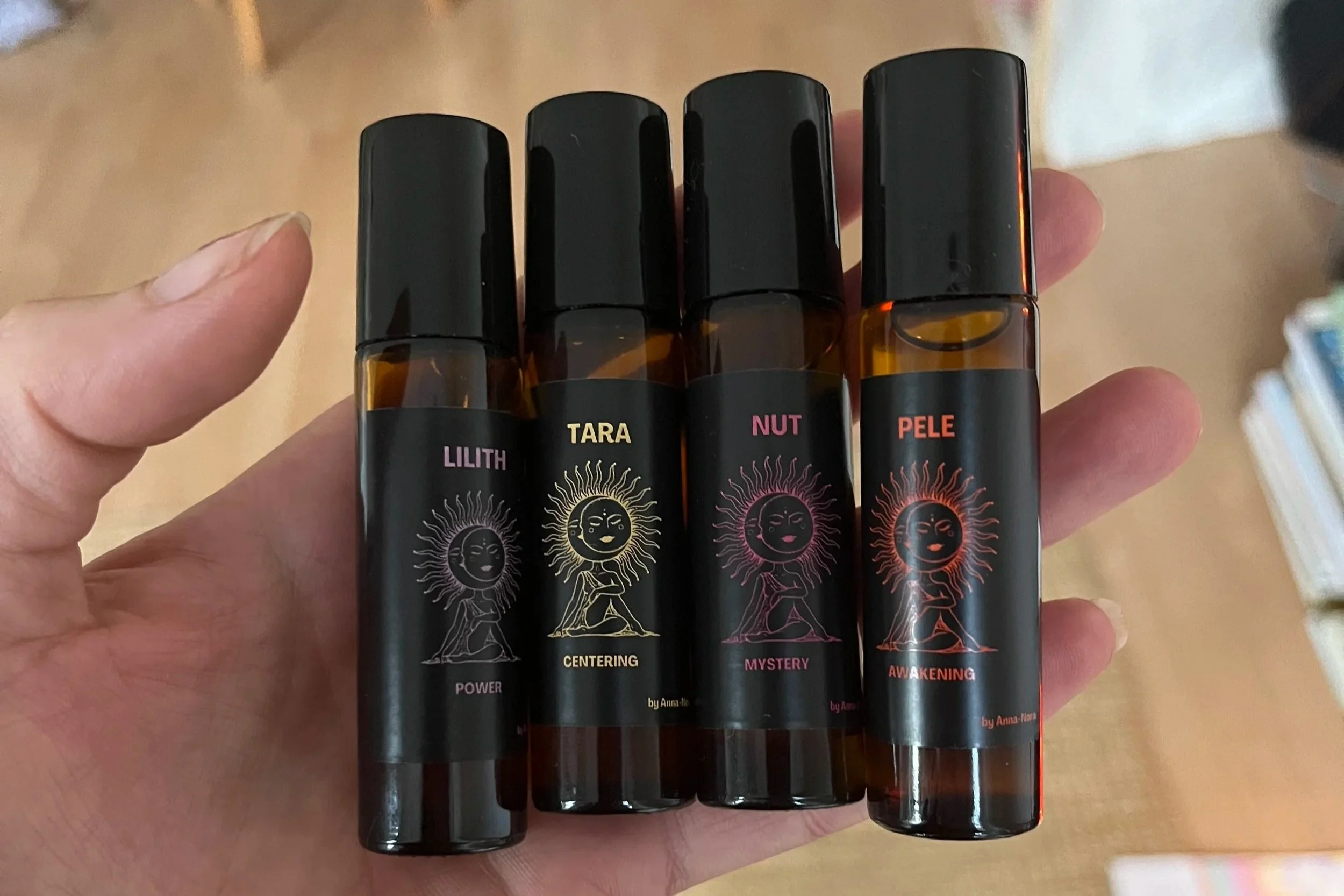A hand holding five black spray bottles with colorful labels and sun illustrations, each labeled with different themes: Lilith, Tara, Nut, Pele, and Awekening.