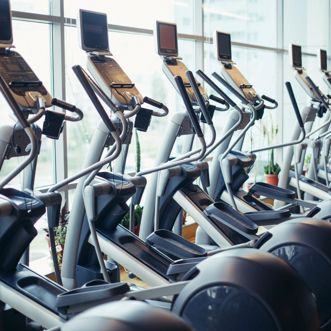 Multiple elliptical exercise machines lined up in a fitness center near large windows, with a cityscape view outside.