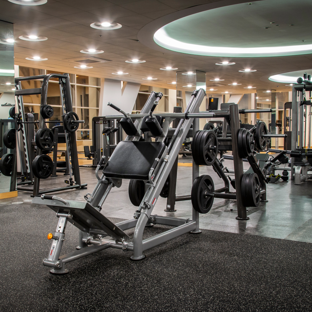 Empty gym with various exercise machines, weight racks, and bright ceiling lights.