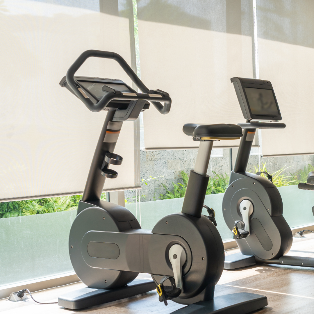 Two stationary exercise bikes with digital screens near large windows with blinds and greenery outside.