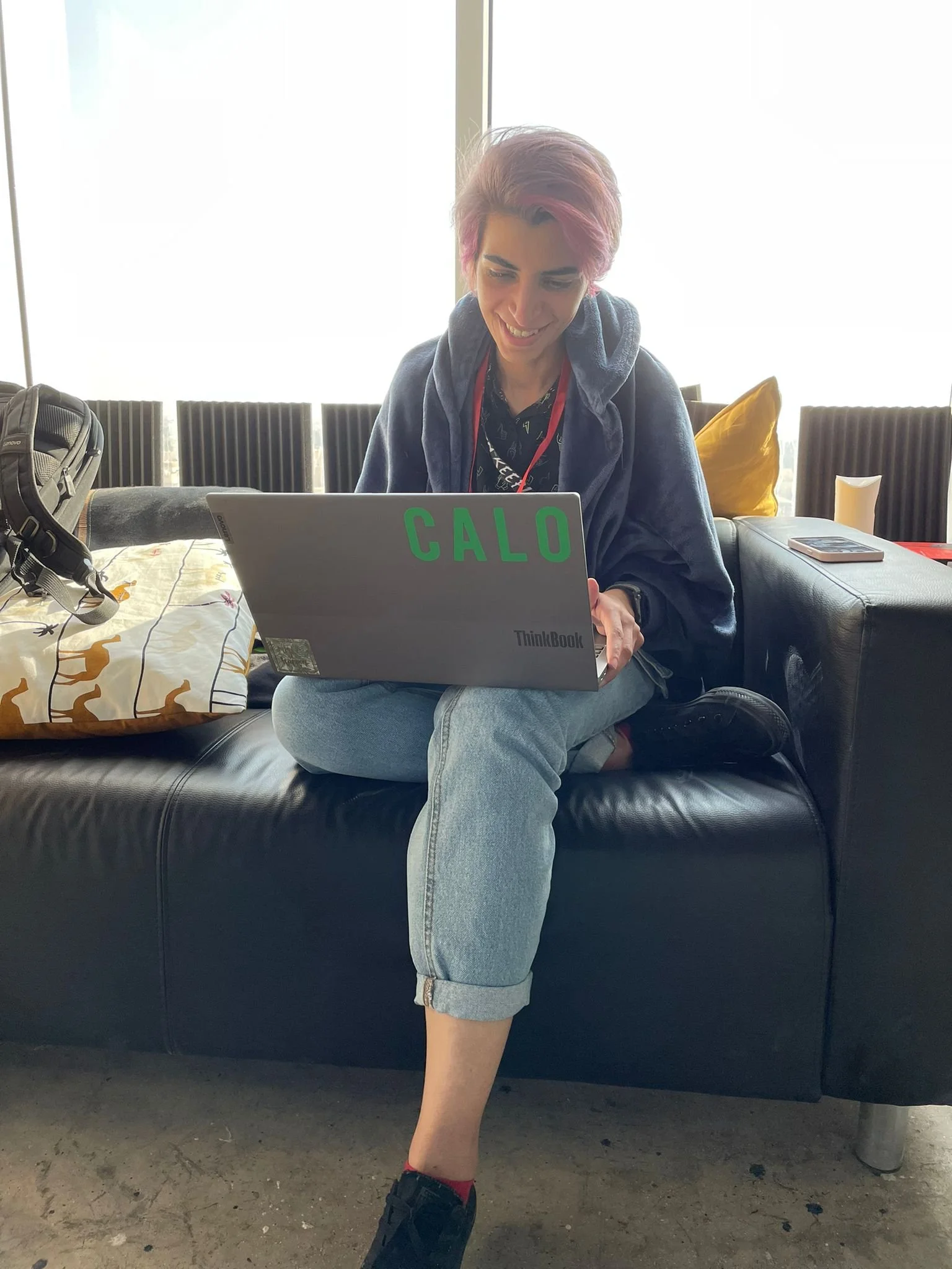 A person with pink hair and a hoodie sitting cross-legged on a black leather couch, working on a silver Lenovo ThinkBook laptop with green letters reading 'CALO' on the lid. There are pillows and a backpack beside them, and a window with daylight in the background.