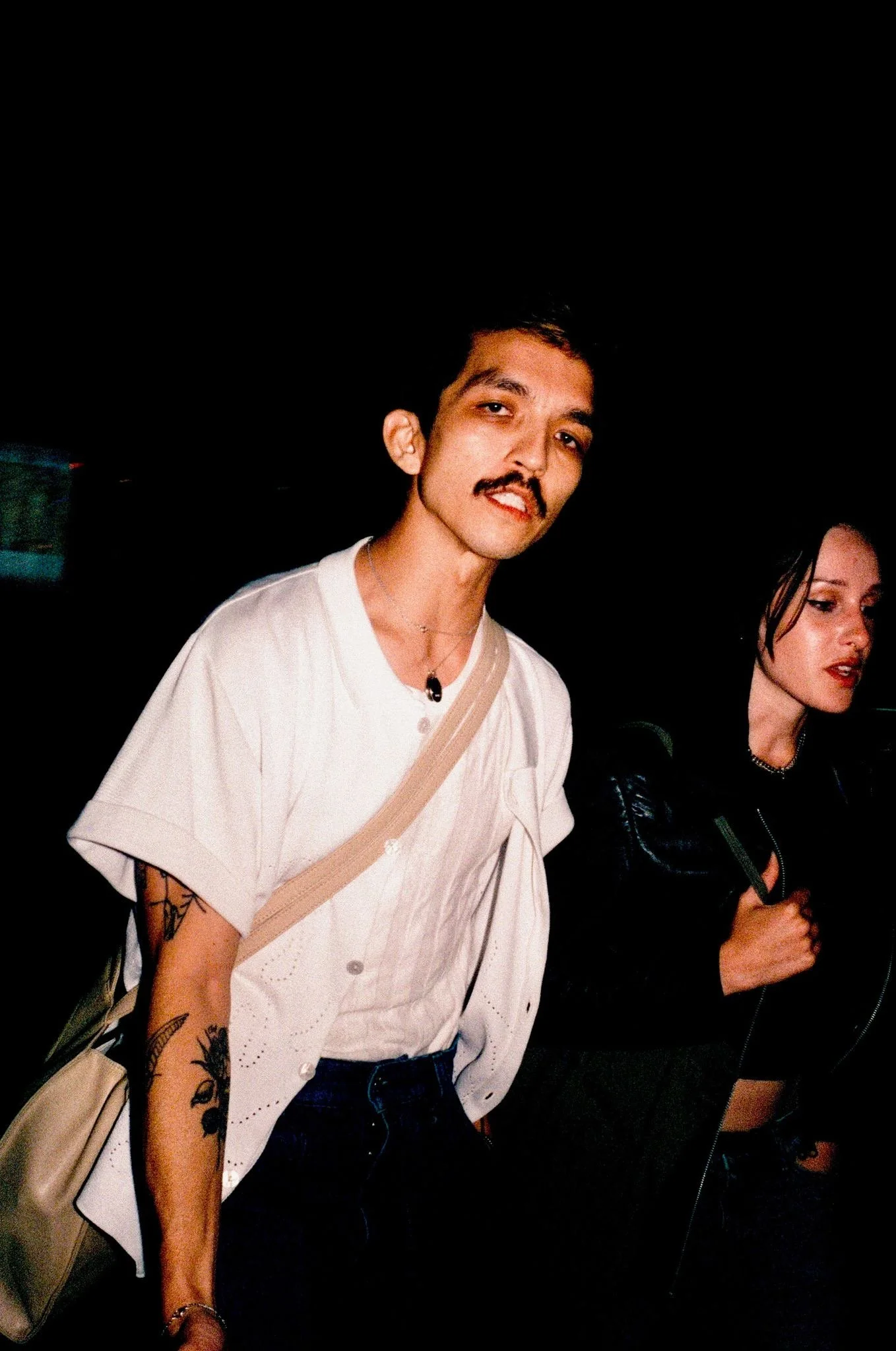 A young man with a mustache and tattoos on his left arm, wearing a white shirt and a beige crossbody bag, standing in a dark environment with a woman beside him, wearing a black leather jacket, dark lipstick, and holding a mobile phone, both appearing to be at a nighttime event or gathering.