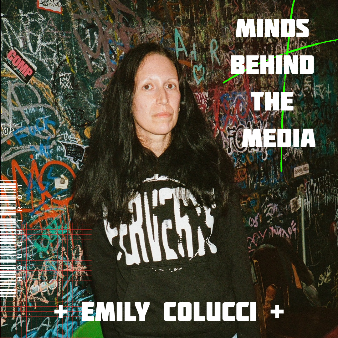 A woman with long dark hair standing in front of a wall covered in colorful graffiti and drawings, wearing a black sweatshirt with white text.