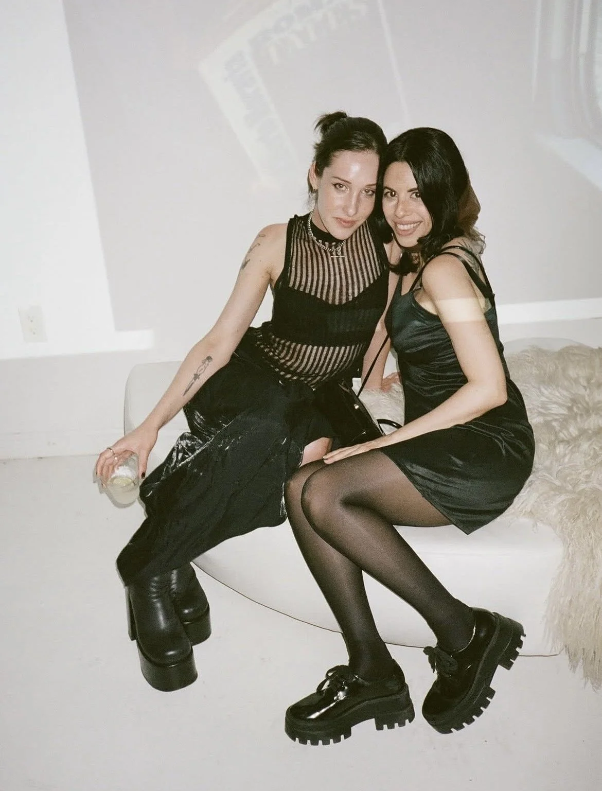 Two women sitting on a white couch in a minimalistic room, wearing black outfits, one holding a drink, smiling and posing for the camera.