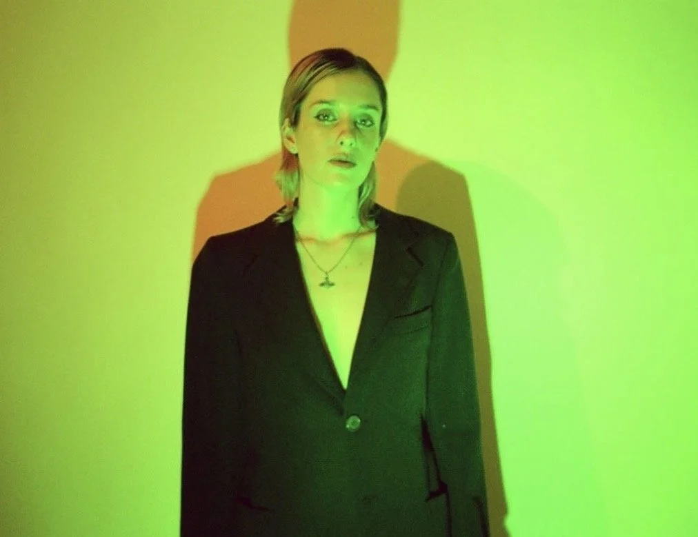 A woman with short blonde hair in a black blazer stands against a green-lit background, casting a shadow on the wall behind her.