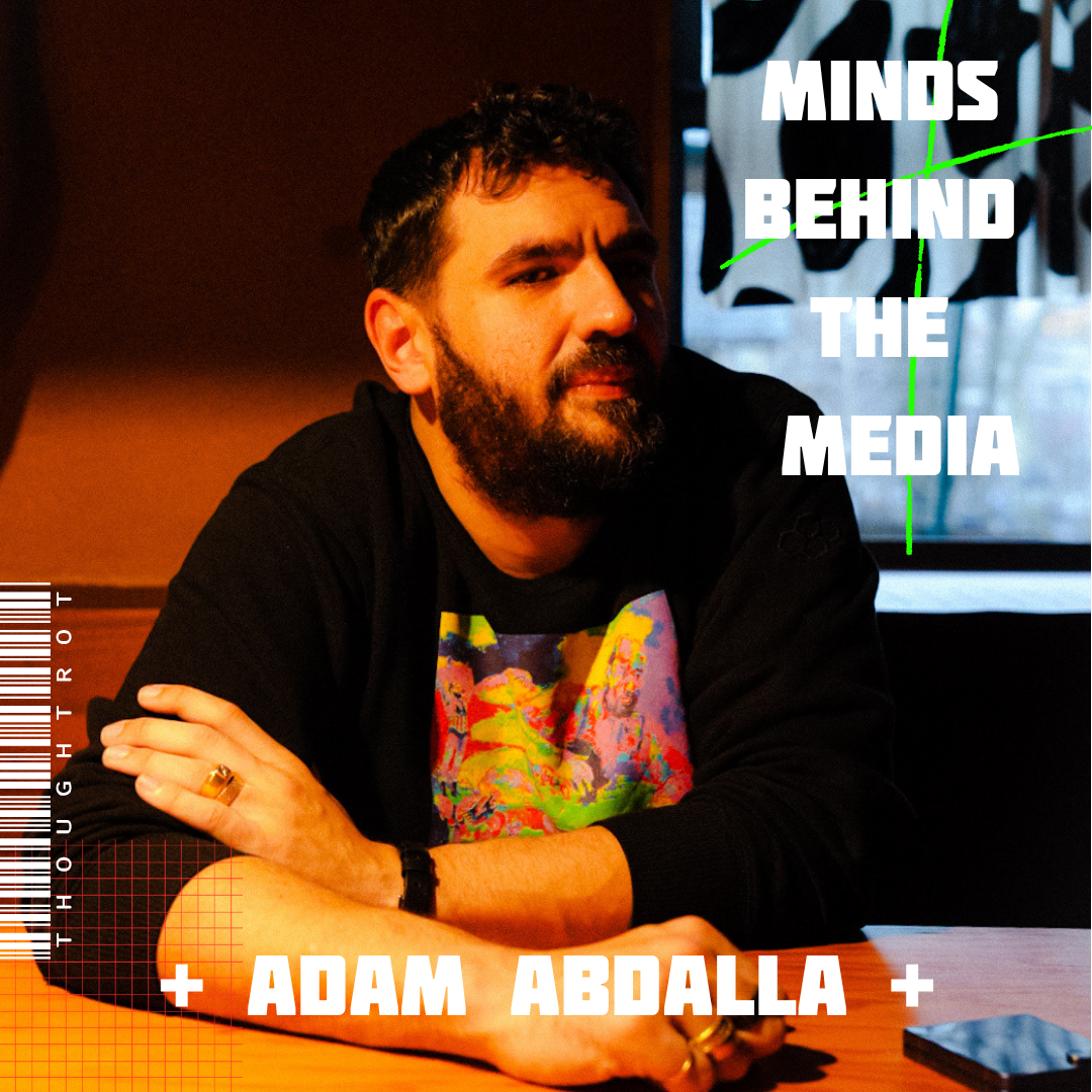 A man with a beard and dark hair sitting at a table with arms crossed, in a dimly lit room, with text overlay that reads 'Minds Behind the Media + Adam Abdalla'.