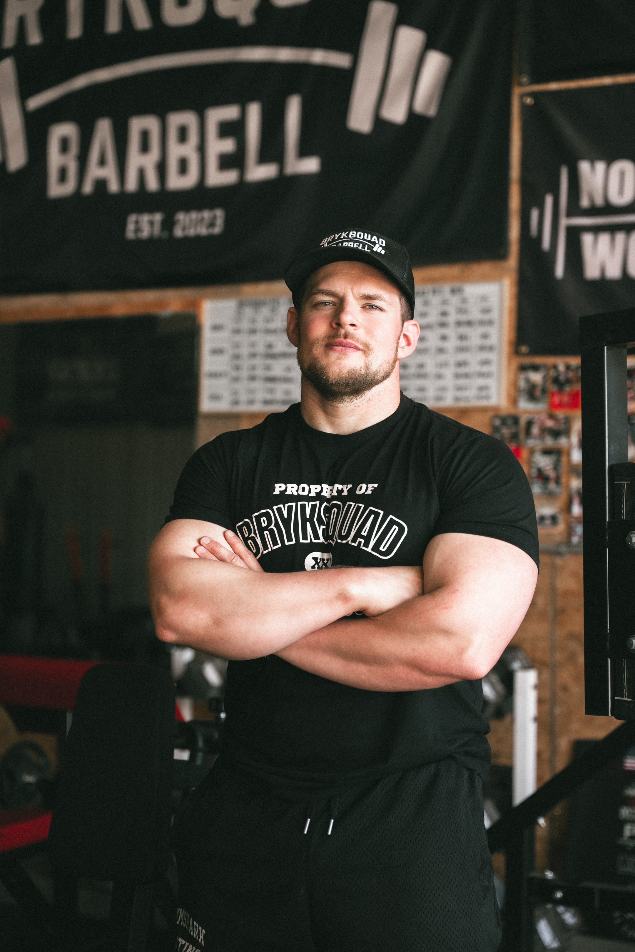 A muscular man in a black T-shirt and cap standing with arms crossed inside a gym, with gym equipment and banners on the walls behind him.