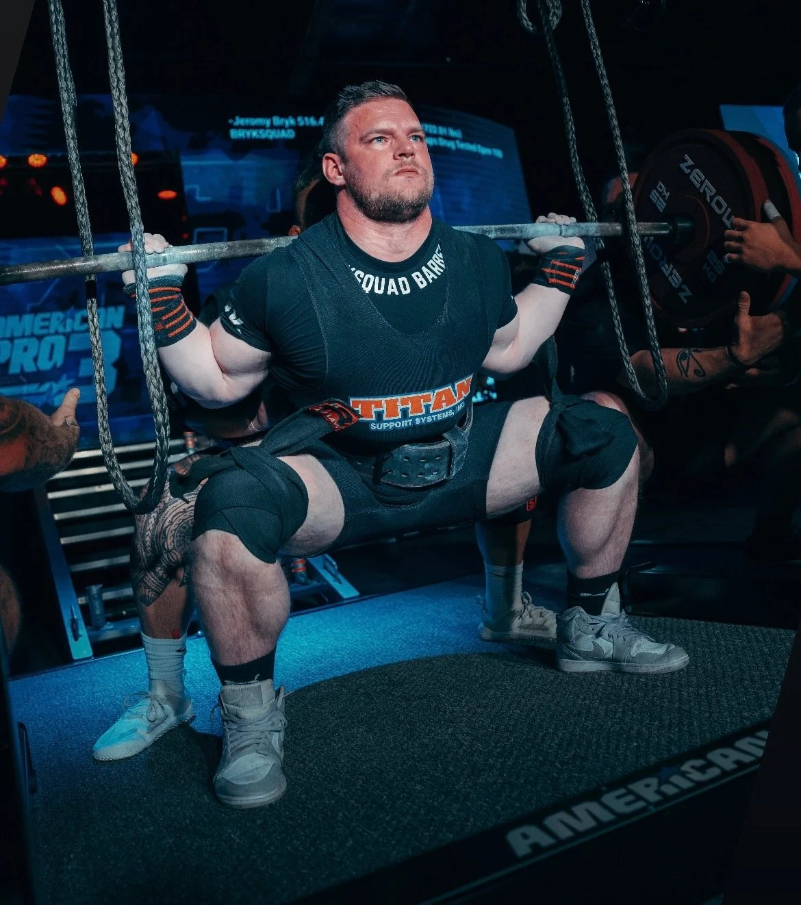 A man performing a squat lift with a barbell at a powerlifting event, wearing a black supportive suit and knee wraps, surrounded by spotters and equipment.