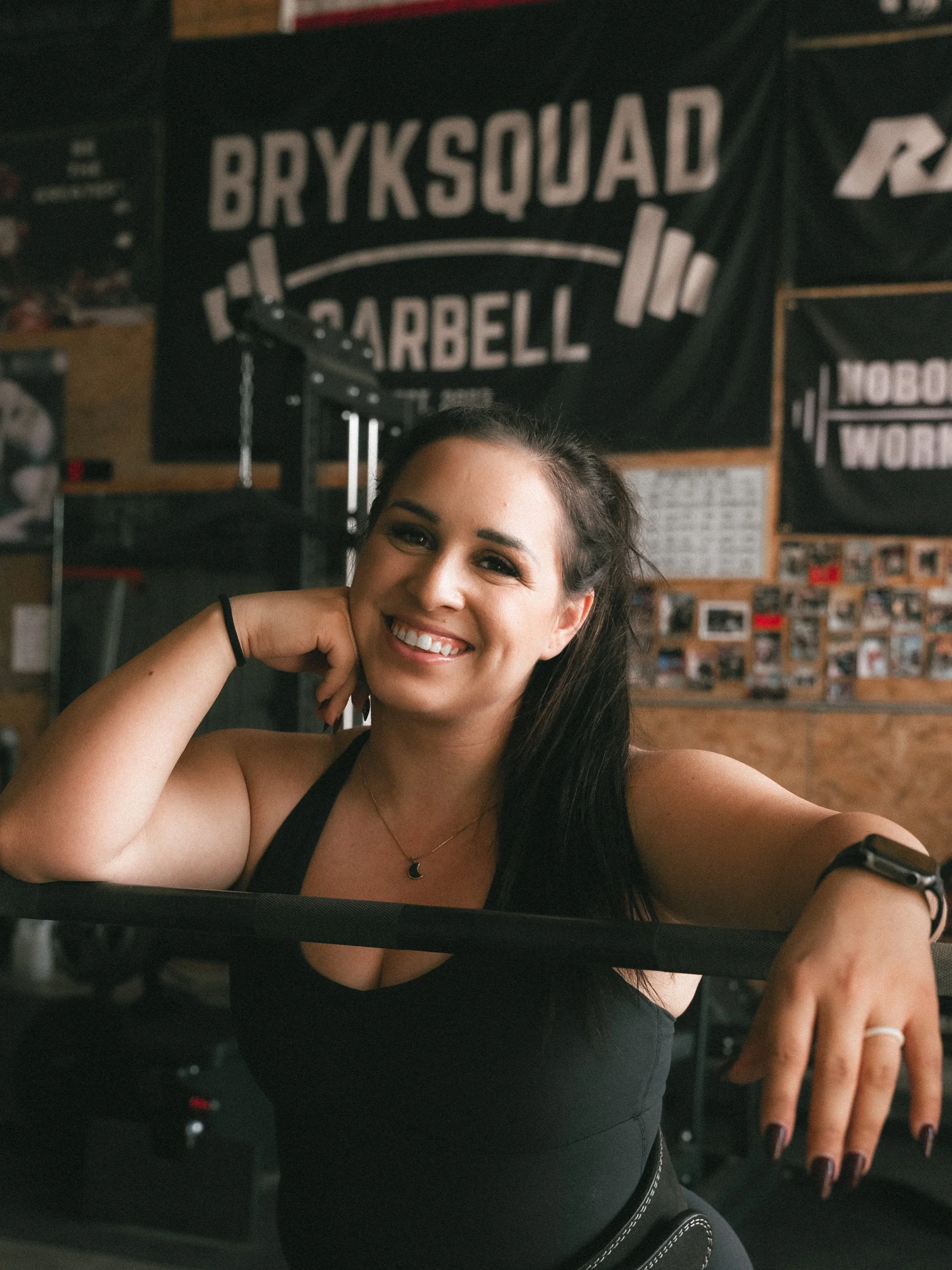 A smiling woman with dark hair in a gym, sitting behind a resistance band, with gym posters and banners on the wall behind her.