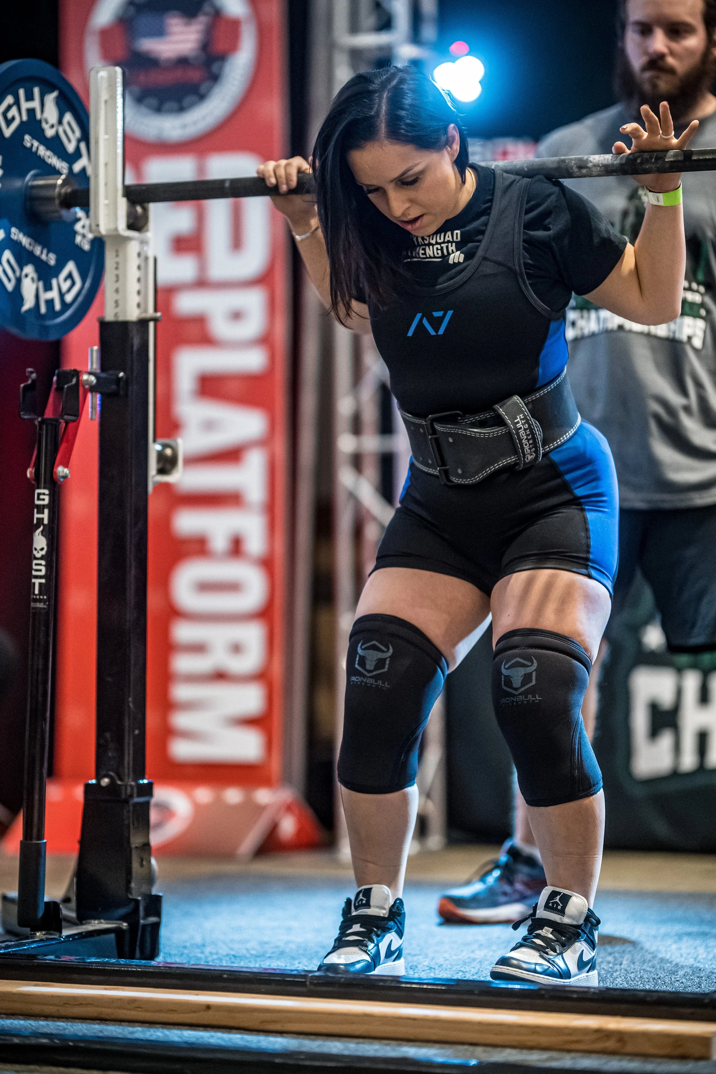 A woman is preparing to lift a barbell during a powerlifting competition, wearing a black and blue singlet, knee sleeves, and weightlifting shoes, with a judge or coach standing nearby.