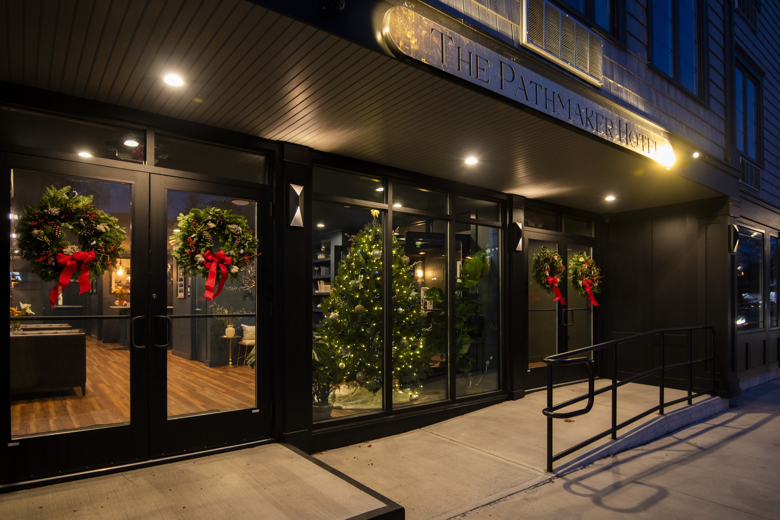 The entrance of a hotel decorated with Christmas wreaths on glass doors and a Christmas tree with lights inside, seen at night.