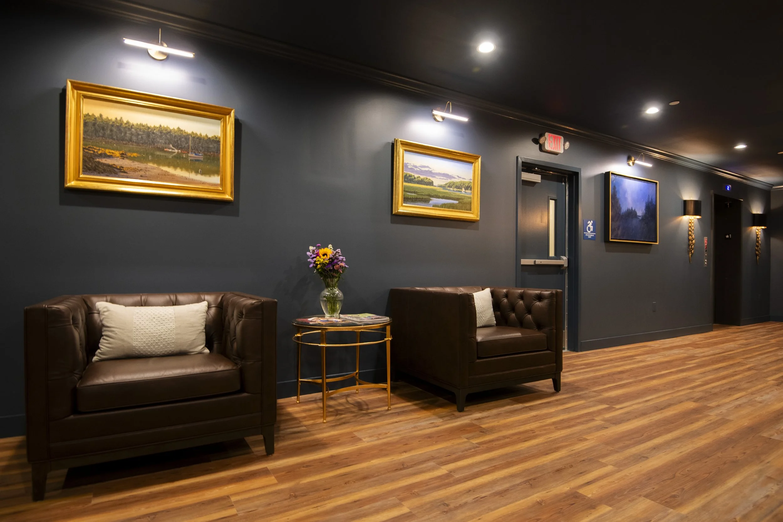 A hotel or waiting area with dark blue walls, wooden flooring, two brown leather armchairs with pillows, a small round side table with a vase of flowers, three landscape paintings on the wall, and an elevator door.