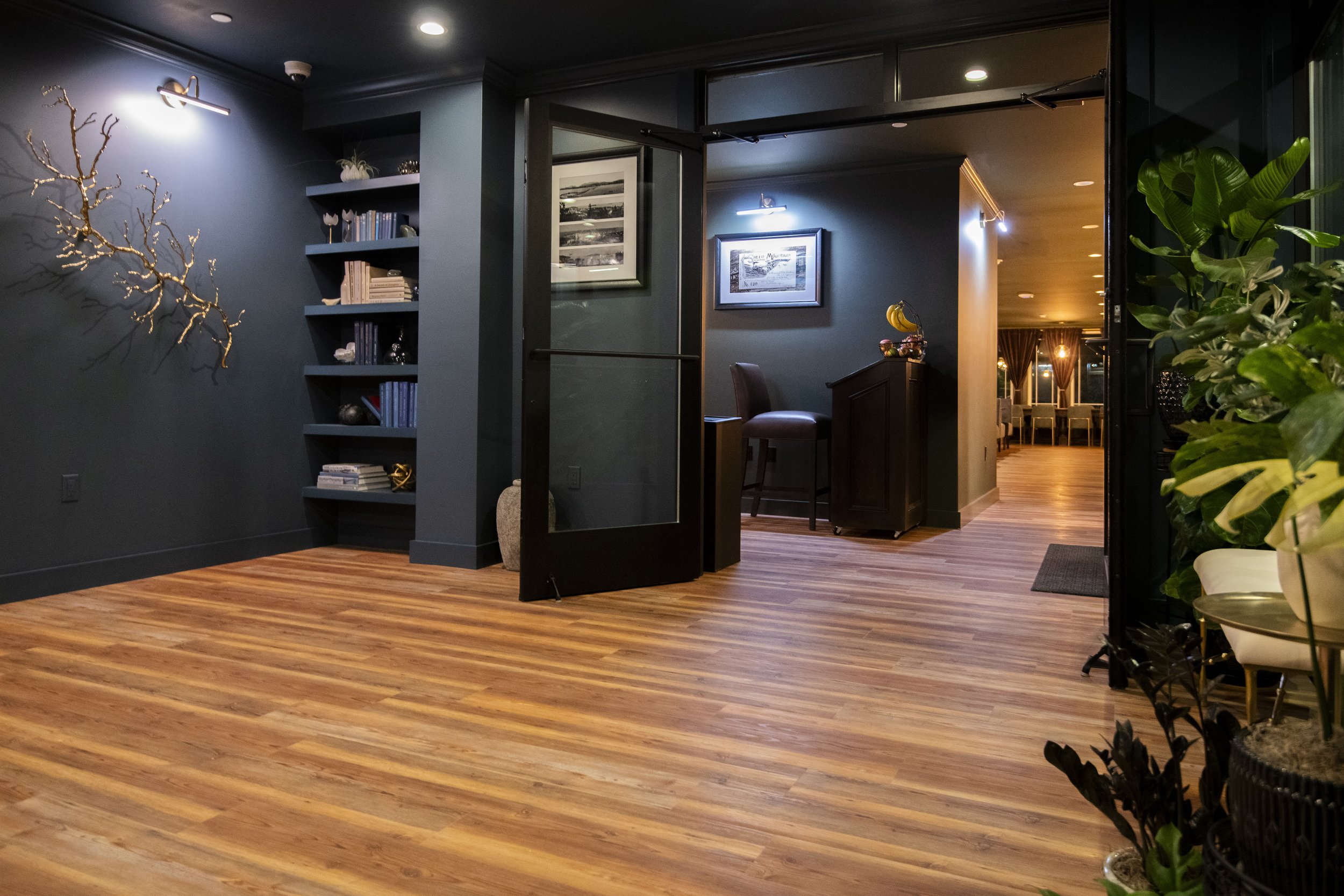 Interior view of a modern lobby or lounge area with dark-colored walls, wooden flooring, a built-in shelf with books and decorative items, wall art, a small bar counter with a stool, potted plants, and a room in the background with dining tables and 