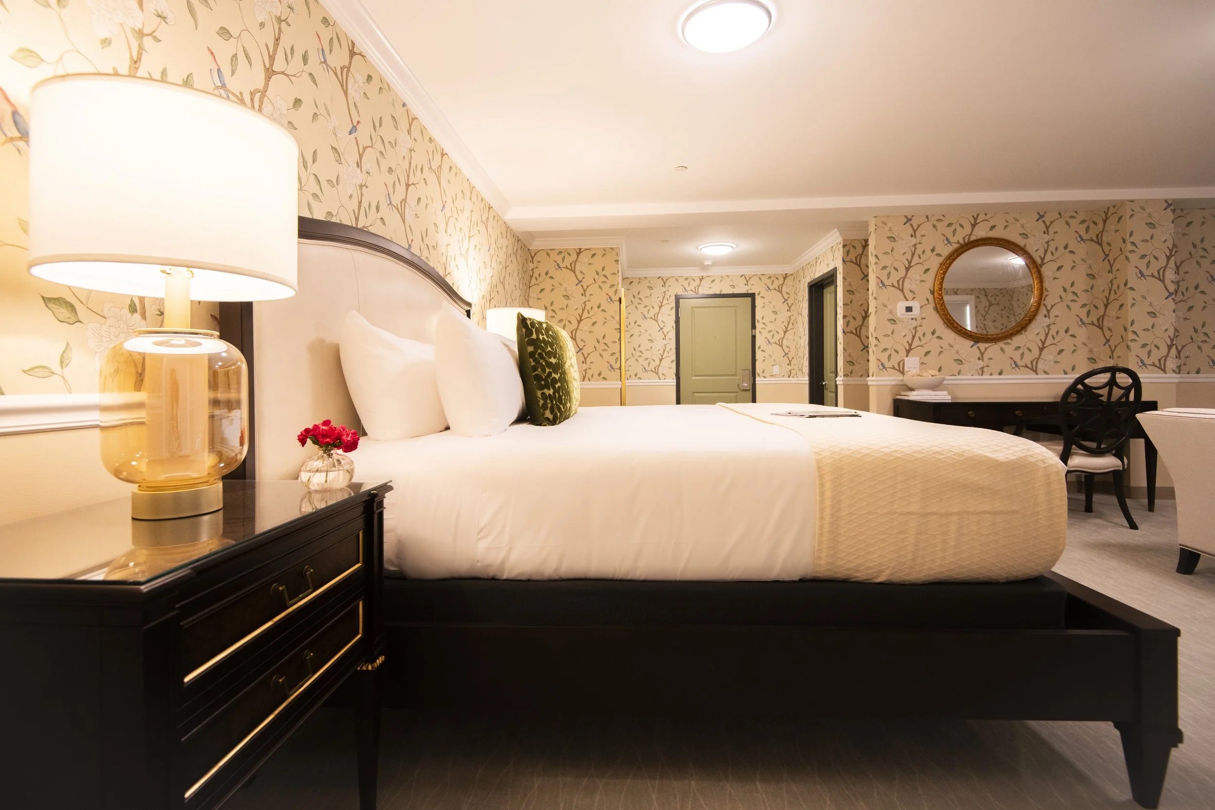 A luxurious hotel bedroom with a large bed, white bedding, and decorative pillows. There is a bedside table with a lamp and a small vase of red flowers. The room has floral wallpaper, a round mirror on the wall, and a desk with a chair. The lighting 