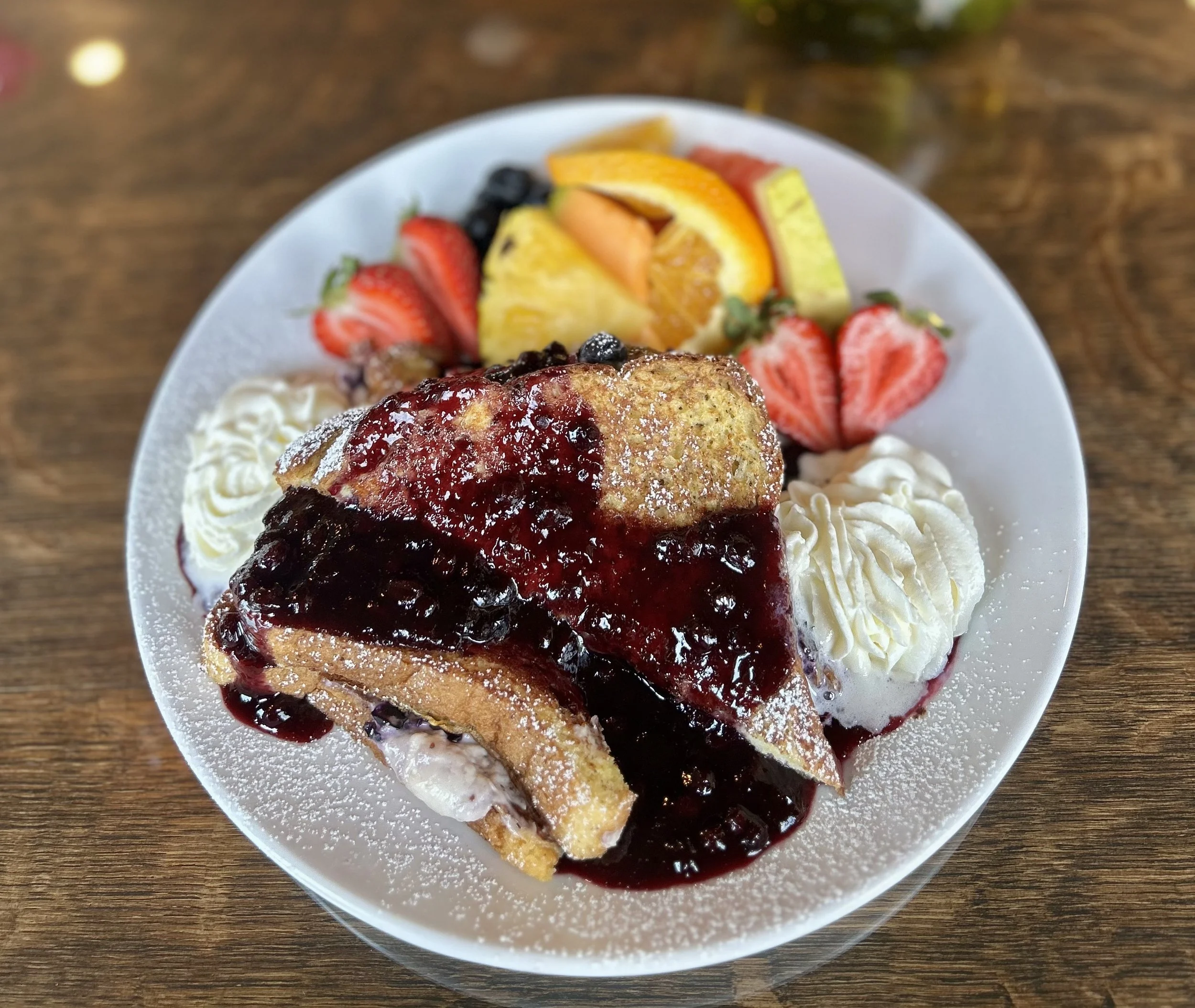 Plate ofFrench Toast topped with blueberry syrup, served with whipped cream, and garnished with fresh strawberries, sliced oranges, and other mixed fruits.