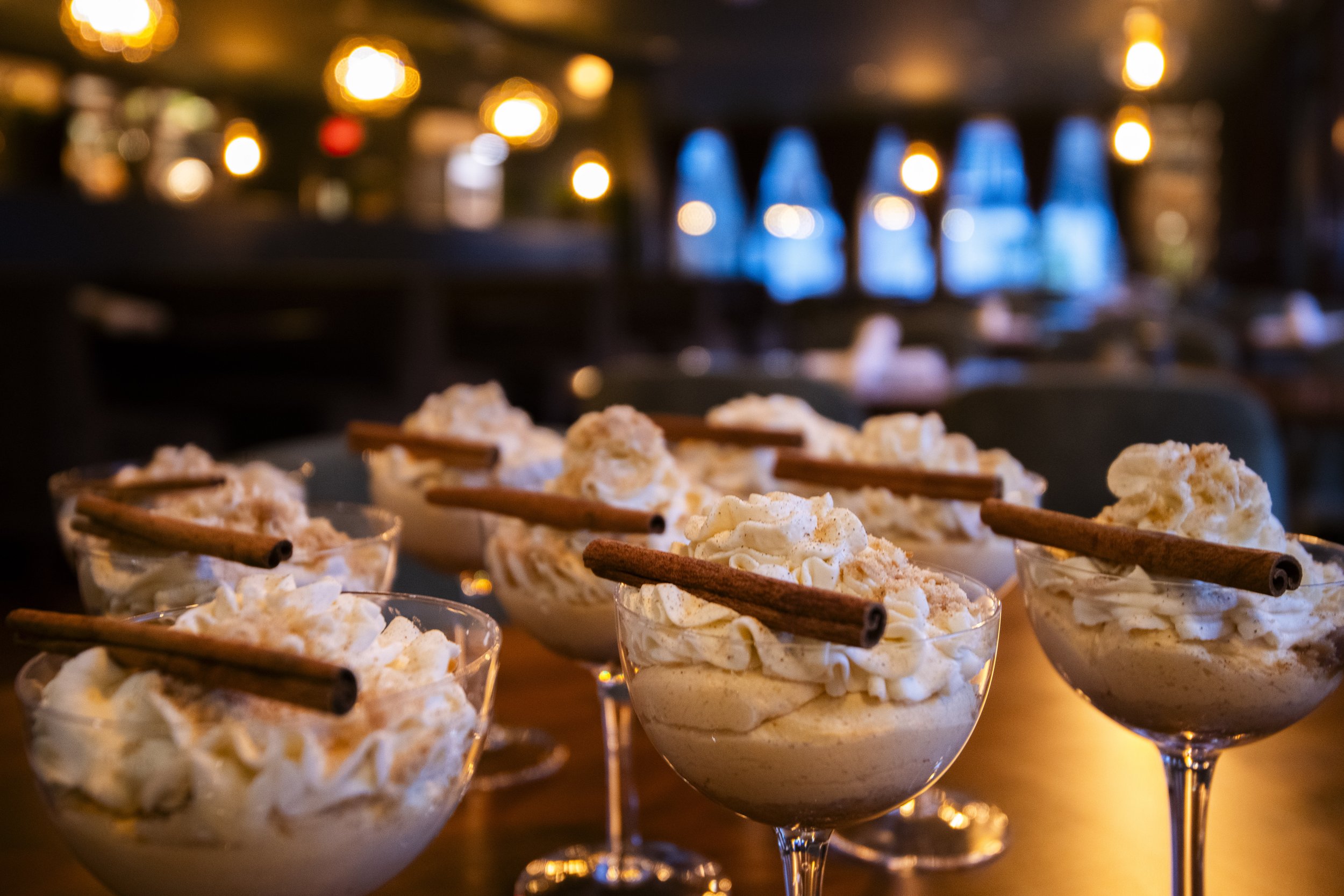 Several glasses filled with whipped cream topped with cinnamon sticks on a wooden surface in a dimly lit indoor setting.