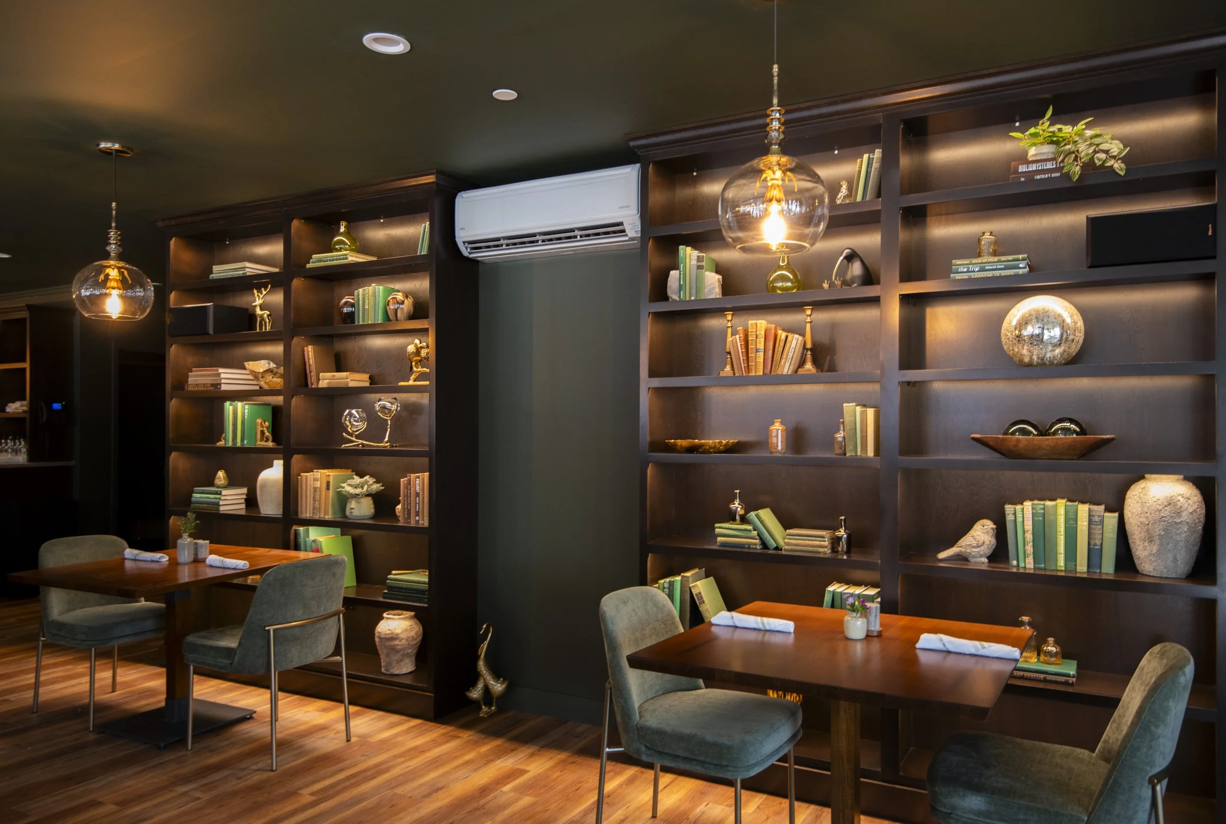 Interior of a cozy restaurant or cafe with dark wood bookshelves filled with decorative items, small potted plants, and books, illuminated by pendant lights and ambient lighting, with wooden tables and plush chairs.