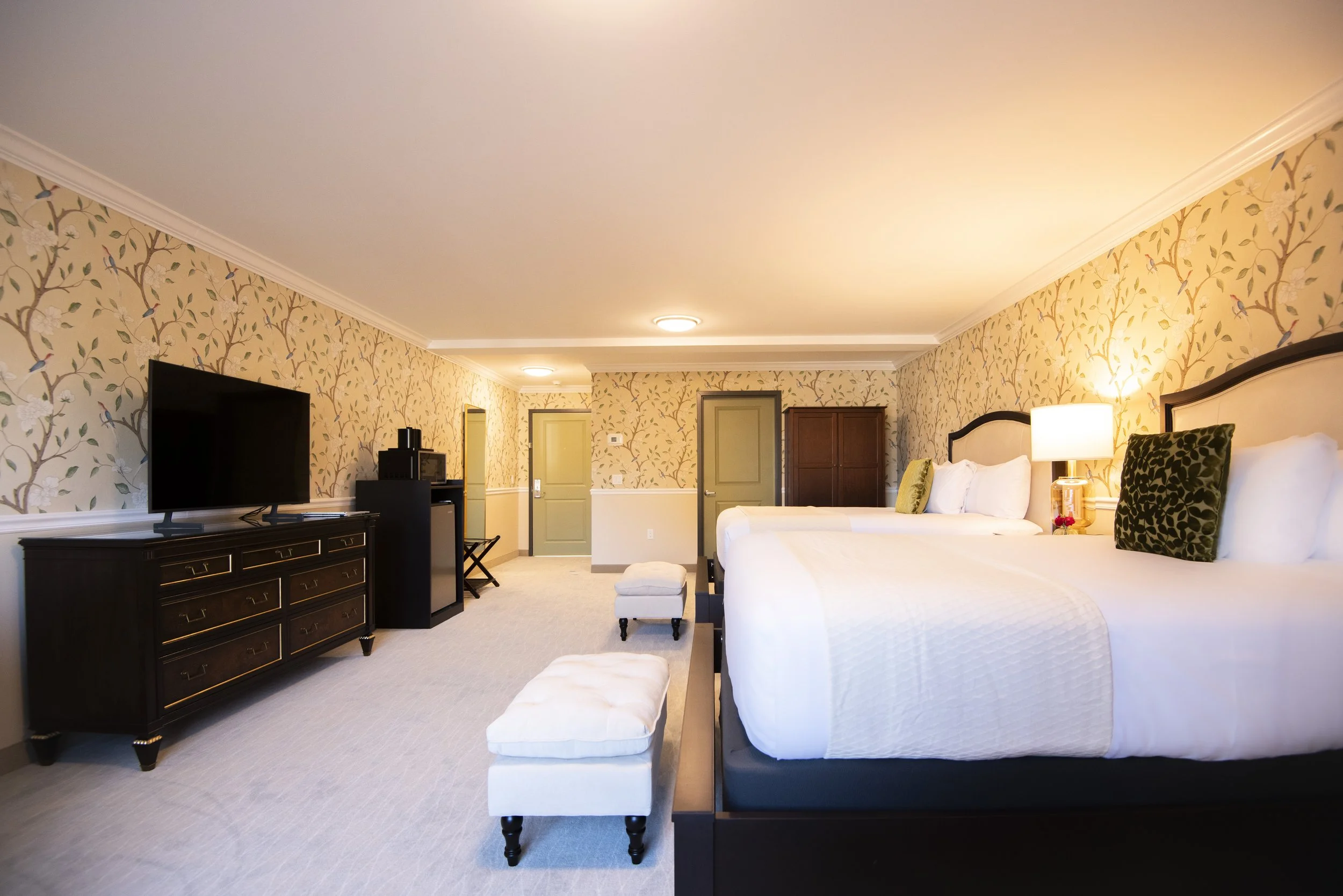 Hotel room with two large beds, a TV on a dark wooden dresser, a small ottoman at the foot of each bed, a lamp on the right bedside table, floral wallpaper, and two doors at the back of the room.