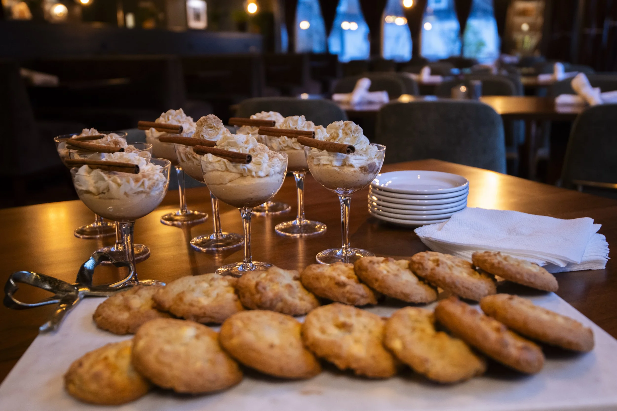 Dessert glasses filled with whipped cream topped with cinnamon stick and cookies on a tray, with additional cookies and plates on a wooden table in a dimly lit restaurant.