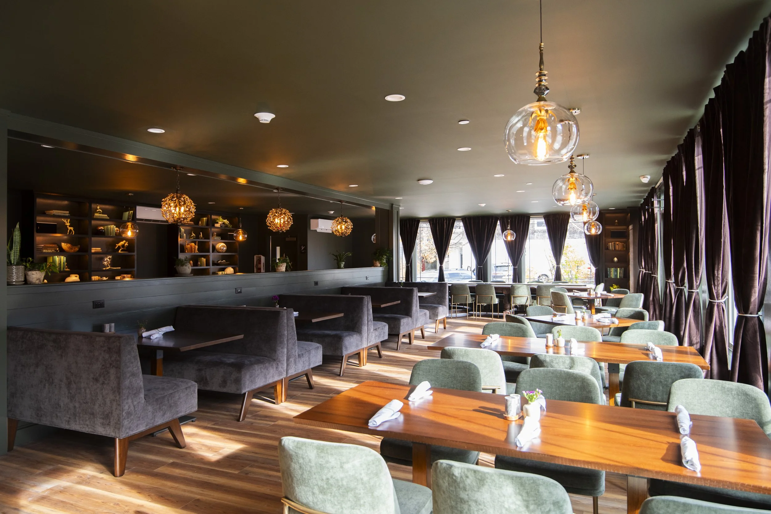 Interior of a modern restaurant with wooden tables, green and gray chairs, large windows with dark curtains, and hanging decorative lights.