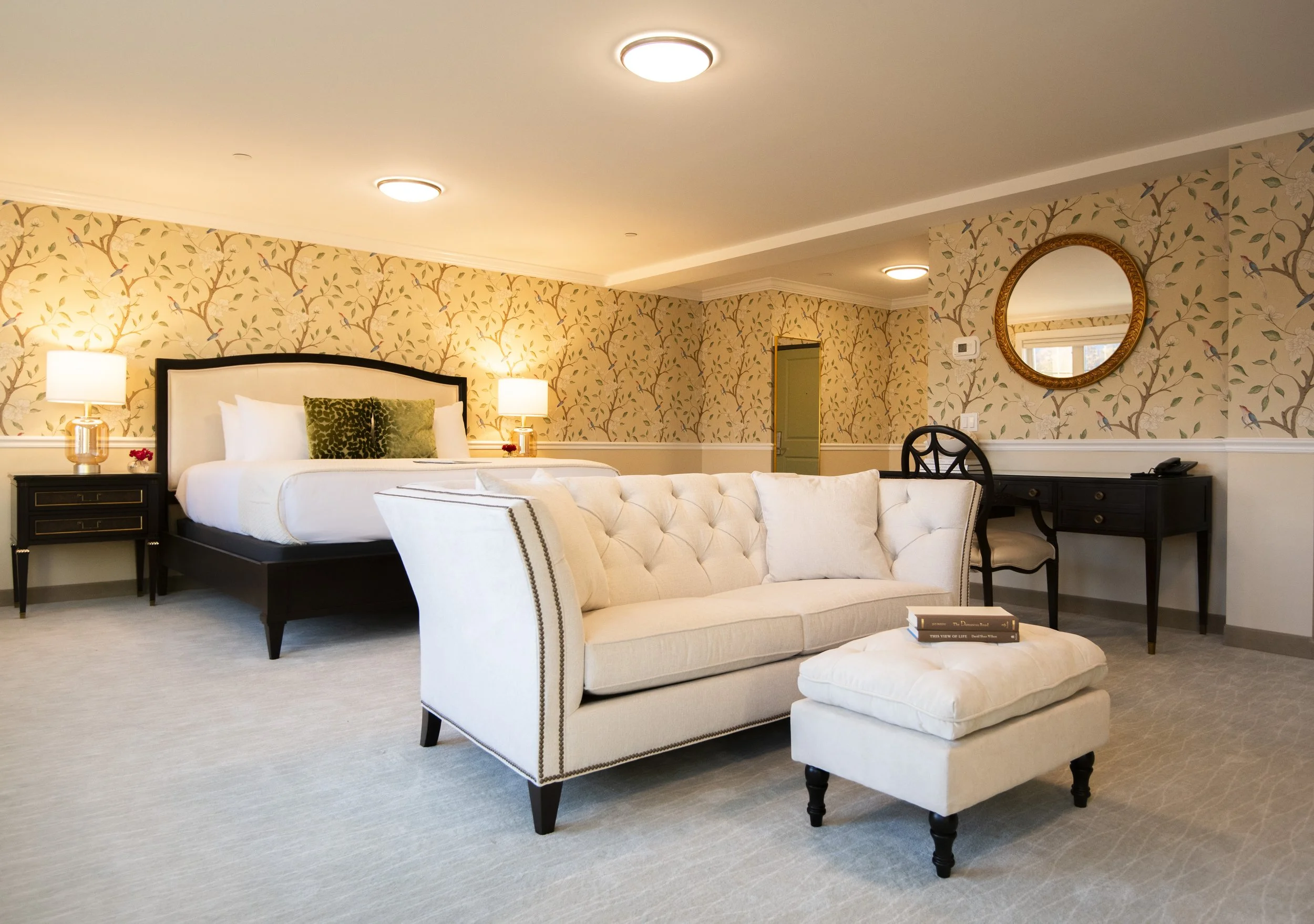 Elegant hotel suite with a white tufted sofa, a bed with white linens, two nightstands with lamps, a black desk with a chair, a round mirror, and floral wallpaper.