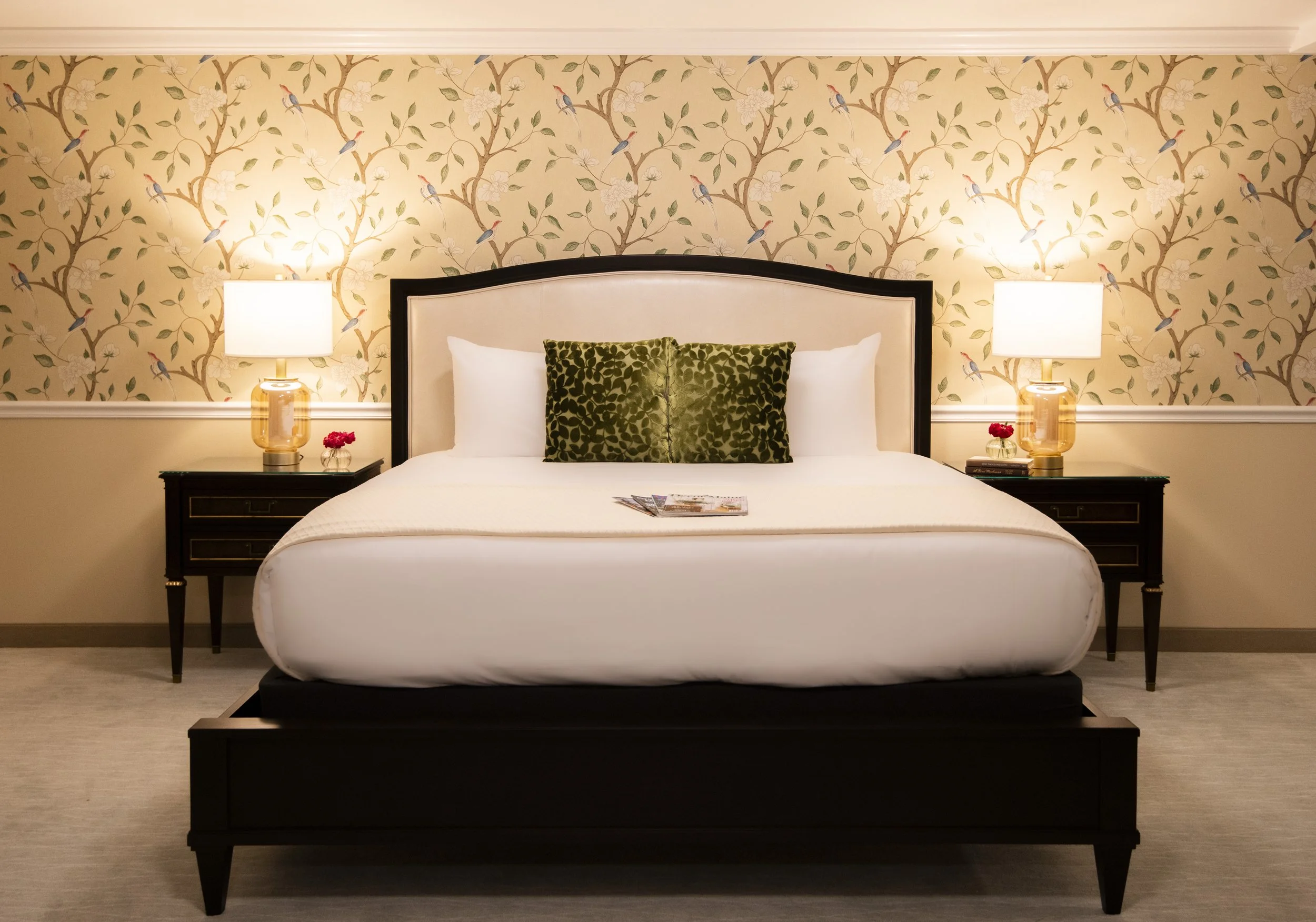 A bedroom with a large bed with a white bedspread, two white pillows, and two green patterned pillows. There are two nightstands with lamps, small vases with red flowers, and magazines on the bed. The wallpaper has a pattern of tree branches with gre